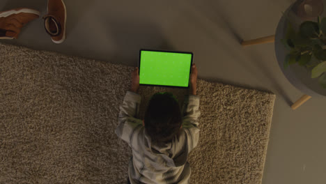 Luftaufnahme-Eines-Kleinen-Jungen,-Der-Zu-Hause-Auf-Einem-Teppich-Liegt-Und-Nachts-Spiele-Spielt-Oder-Auf-Ein-Digitales-Tablet-Mit-Grünem-Bildschirm-Streamt-1